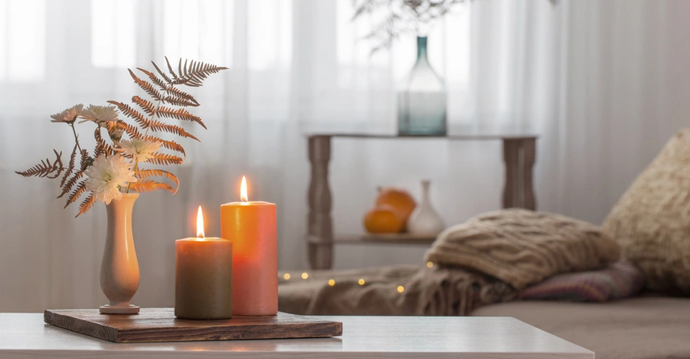 zwei brennende Stumpenerzen in orange und beige stehen auf Holzbrett auf Couchtisch, im Hintergrund eine Couch mit Kissen und Decke. Herbstliche, gemütliche Stimmung
