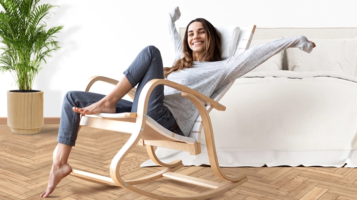 Galerie-Bild 1: Eine lächelnde Frau sitzt entspannt in einem hellen Schlafzimmer auf einem geschwungenen Holz-Schaukelstuhl. Sie streckt die Arme aus, trägt ein graues Langarmshirt und dunkle Hose. Im Hintergrund stehen ein großes Bett mit heller Bettwäsche und eine grüne Zimmerpflanze auf einem Parkettboden.