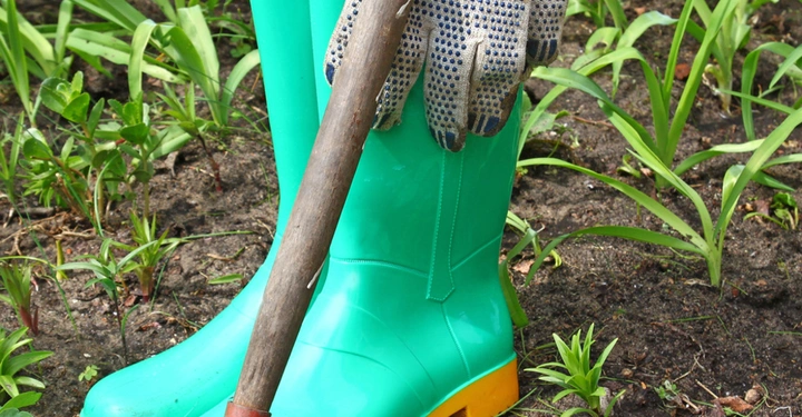 Grüne Gummistiefel mit Handschuhen darauf stehen auf Erde neben einer Hacke im Gartenumfeld mit Pflanzen und Unkraut.