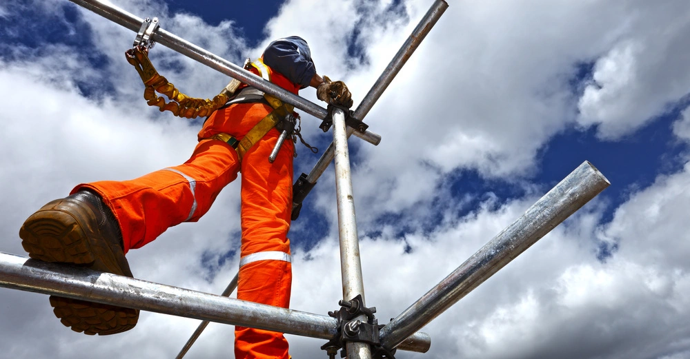 Arbeiter in orangefarbener Schutzkleidung klettert ein Gerüst hoch in einer Höhe gegen einen wolkigen Himmel.