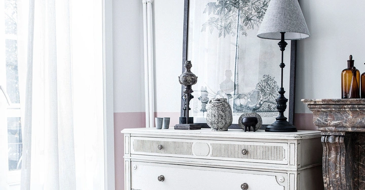 Weißer Schrank steht still im hell erleuchteten Raum dekoriert mit einer Lampe einer Vase und gerahmter botanischer Kunst auf einer Marmorplatte neben einem Fenster mit durchsichtigem Vorhang