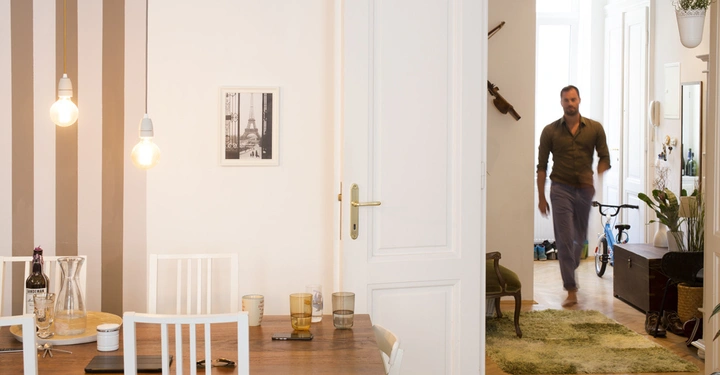 Galerie-Bild 3: Mann geht in einem Flur einer Wohnung Hintergrund Esszimmer mit Holztisch vier Stühlen zwei hängenden Glühbirnen Flaschen und Gläsern und Bild an der Wand