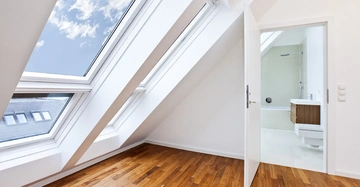 Schräge Dachfenster lassen Sonnenlicht in ein Zimmer mit Holzboden und weißen Wänden einfallen. Eine geöffnete Tür zeigt ein weißes Badezimmer mit Duschkabine und Holzwaschbecken.