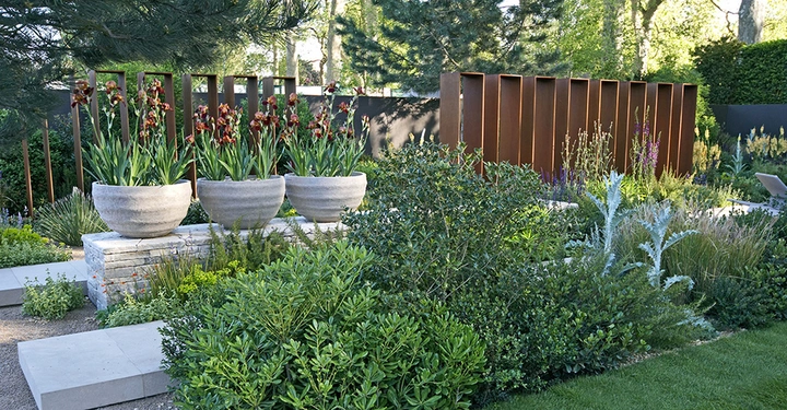 Große Betonschalen mit roten Blumen auf erhöhtem Steinpodest Stehende Metallplatten und üppige grüne Vegetation im Hintergrund Moderner Gartenumgebung