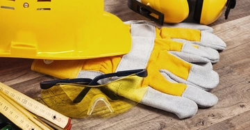 Gelber Schutzhelm Handschuhe Schutzbrille und Gehörschutz auf einem Holztisch. Neben dem Helm liegen ein Messband und eine Wasserwaage.