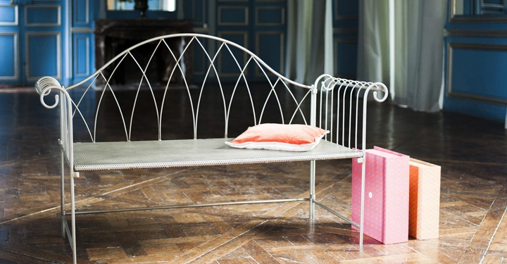 Galerie-Bild 6: Metallbank mit einem orangefarbenen Kissen darauf daneben zwei farbige Kartons in einem Raum mit Holzboden und blauen Wandvertäfelungen. Im Hintergrund ein Tisch mit Blumenarrangement.