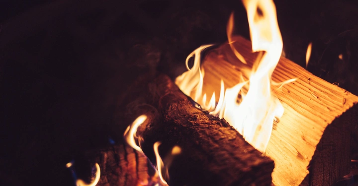 Brennende Holzscheite mit lodernden Flammen in Nahaufnahme. Der Hintergrund ist dunkel und im Schatten gehalten, was die Helligkeit des Feuers betont.