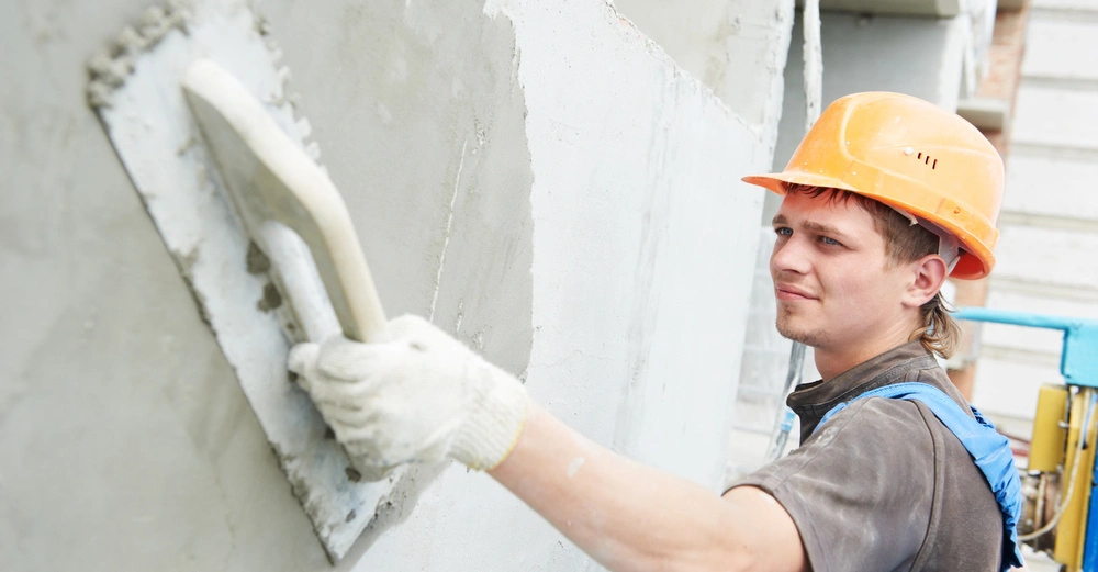 Arbeiter trägt eine orangefarbene Schutzhelm und blaue Arbeitskleidung er glättet mit einer Kelle eine Wand bei einer Baustelle im Freien
