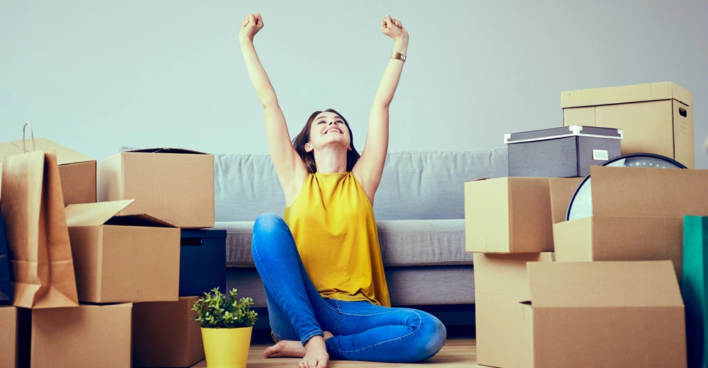 Frau sitzt jubelnd auf dem Boden umgeben von Umzugskartons und einer kleinen Pflanze vor einem grauen Sofa in einem Wohnzimmer.