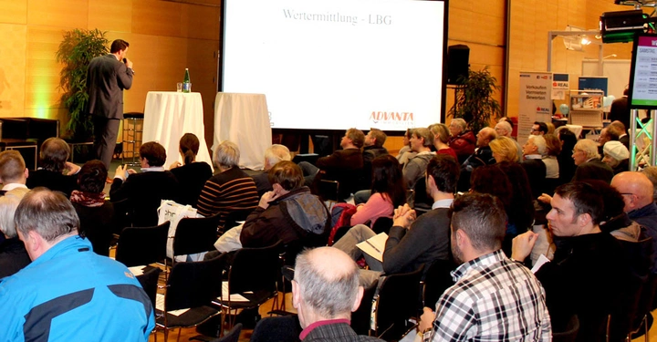 Galerie-Bild 4: Ein Mann hält einen Vortrag auf einer Bühne vor einem sitzenden Publikum in einem Konferenzraum. Auf der großen Leinwand steht Wertermittlung - LBG ADVANTA.