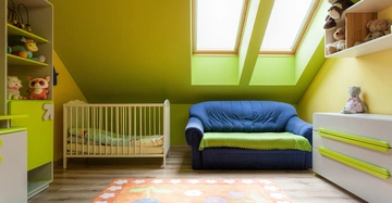 Ein Babybett steht in einem gelbgrünen Zimmer neben einem blauen Sofa unter Dachfenstern Spielzeugregale und eine Kommode umgeben das Sofa orangener Teppich mit Blumenmuster im Vordergrund