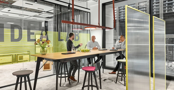Galerie-Bild 1: Drei Männer sitzen und unterhalten sich an einem hohen Tisch in einem modernen Büro mit großen Fenstern. Im Hintergrund steht auf einer Glaswand der Text IDEA LAB.