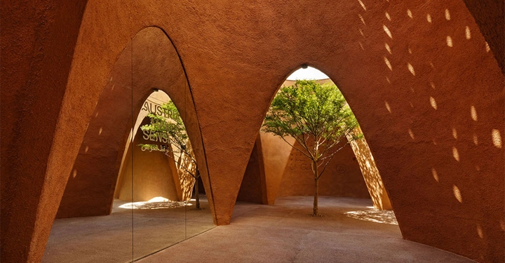 Galerie-Bild 2: Baum steht inmitten einer architektonischen Struktur mit bronzefarbenen, gewölbten Wänden, die von natürlichen Lichtmustern durchbrochen werden; Schriftzug "Susteren Sens Seni" in verschiedenen Sprachen auf einer Glastür