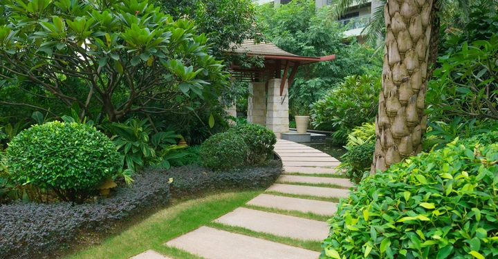 Ein Gartenweg führt durch üppige grüne Vegetation zu einem kleinen Pavillon mit einem Ziegeldach in einem friedlichen Gartenumfeld.