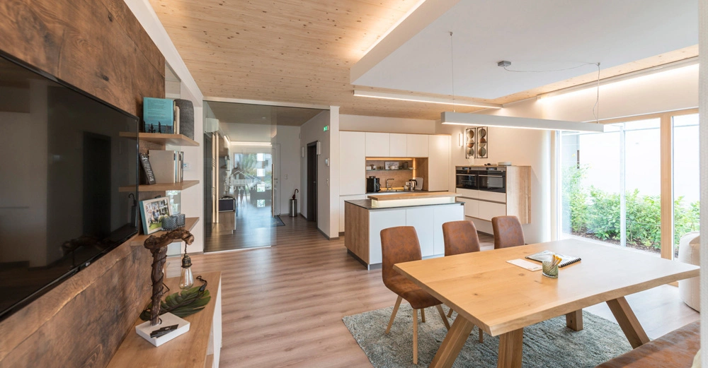 Holztisch und vier Stühle stehen im Vordergrund in einem modernen Esszimmer mit Holzboden und -decke im Hintergrund offene Küche und Wohnzimmer mit Dekorationen und Bücherregalen auf hölzerner TV-Wand.