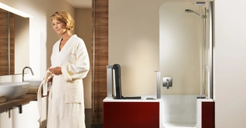 Eine Frau in einem weißen Bademantel steht vor dem Waschbecken in einem modernen Badezimmer mit einer integrierten Badewanne und Dusche vor einer holzverkleideten Wand. Text: keine.