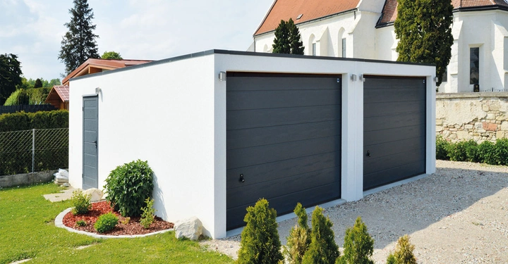 Weiße Doppelgarage mit grauen Toren und Tür steht auf einem Kiesplatz vor einem Garten und Haus mit rotem Dach im Hintergrund. Bäume und Sträucher säumen den Rasen.