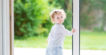 Kleinkind schaut nach hinten hält Tür auf hellem Boden im Innenbereich dahinter verschwommene grüne Gartenlandschaft.