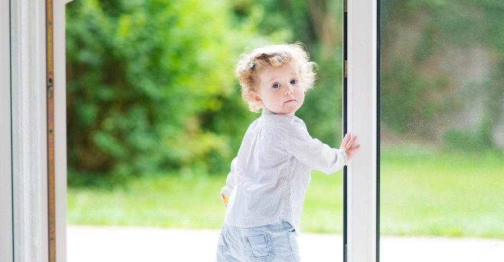 Kleinkind schaut nach hinten hält Tür auf hellem Boden im Innenbereich dahinter verschwommene grüne Gartenlandschaft.