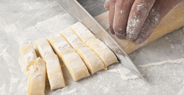 Eine Hand schneidet mit einem Messer einen länglichen Teig in gleichmäßige Stücke auf einer bemehlten Arbeitsfläche.