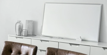 Weiße Tafel ruht auf einem weißen Schrank in einem modernen Büroraum mit Dekorationen wie Vasen und Kerzen. Davor sind zwei braune Ledersessel.