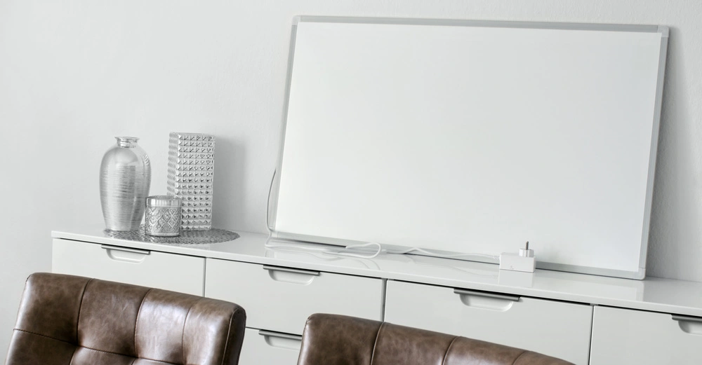 Weiße Tafel ruht auf einem weißen Schrank in einem modernen Büroraum mit Dekorationen wie Vasen und Kerzen. Davor sind zwei braune Ledersessel.