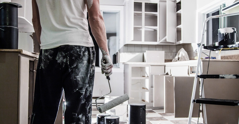 Person streicht mit Farbroller eine Kücheneinrichtung in einem Raum mit halb leeren Schränken und mehreren Farbdosen auf dem Boden