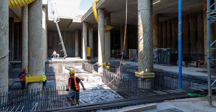 Galerie-Bild 6: Mehrere Bauarbeiter arbeiten an einem Gebäude mit großen Betonpfeilern. Sie tragen orangefarbene Westen und Helme. Im Hintergrund sind verschiedene Baugerüste und Materialien zu sehen.