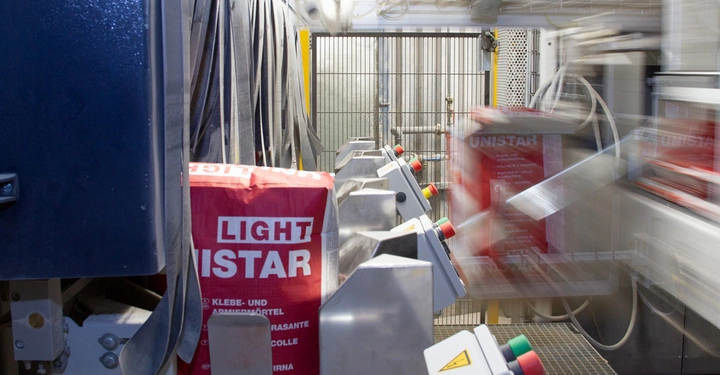Rote UNISTAR LIGHT Säcke bewegen sich auf einem Fließband in einer industriellen Verpackungsmaschine. Metallische Hebel und Steuerknöpfe umgeben die Säcke in einem hellen, maschinellen Raum. Text auf den Säcken: UNISTAR LIGHT KLEBE- UND ARMIERMörtEL