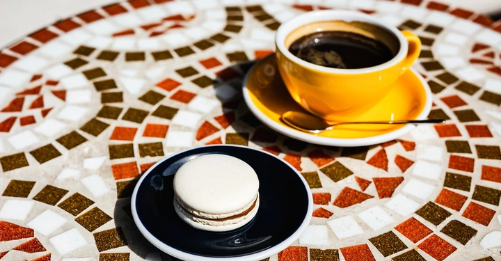 Galerie-Bild 4: Runder Mosaiktisch mit weißen, rostroten und braunen Steinchen. Darauf eine dottergelbe Kaffetasse und ein schwarzes Tellerchen mit einem Macaron
