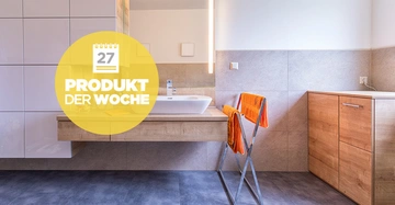 Ein hölzerner Waschtisch mit Waschbecken in einem modernen Badezimmer, Handtücher auf einem Handtuchhalter, daneben ein Holzschrank. Text: 27 PRODUKT DER WOCHE.
