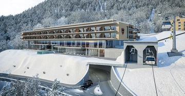 Großes modernes Hotel steht auf einem verschneiten Hügel Gäste sitzen auf Balkonen und im Innenbereich im Hintergrund gibt es schneebedeckte Bäume Seilbahn führt vom Hotel weg Aufschrift RAM