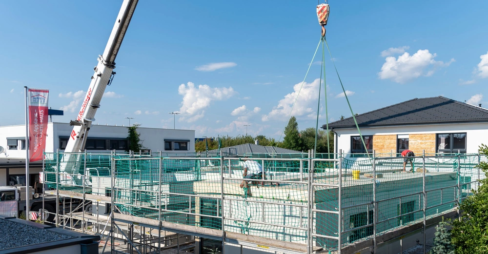 Ein Kran hebt Baumaterialien auf ein im Bau befindliches Gebäude Arbeiter sind auf dem Dach beschäftigt Im Hintergrund befinden sich moderne Wohnhäuser und der Himmel ist blau mit vereinzelten Wolken Text: Applaus Applaus hartl HAUS