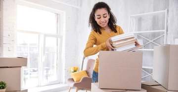 Eine Frau packt Bücher in einen Karton in einem hellen Raum mit Umzugskartons einem modernen Stuhl und einem leeren Regal im Hintergrund