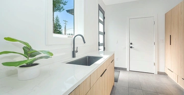 Weiße Küchenspüle mit schwarzer Armatur und Topfpflanze auf hellgrauer Arbeitsplatte, modernes Hausinterieur mit Holzmöbeln und großen Fenstern, das Tageslicht von außen hereinlässt.