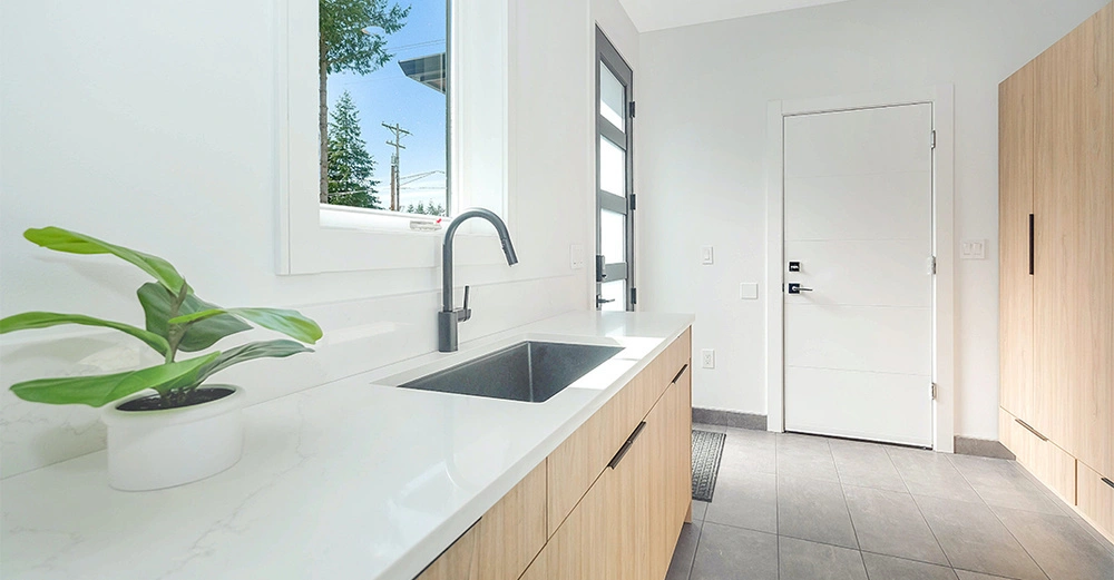 Weiße Küchenspüle mit schwarzer Armatur und Topfpflanze auf hellgrauer Arbeitsplatte, modernes Hausinterieur mit Holzmöbeln und großen Fenstern, das Tageslicht von außen hereinlässt.