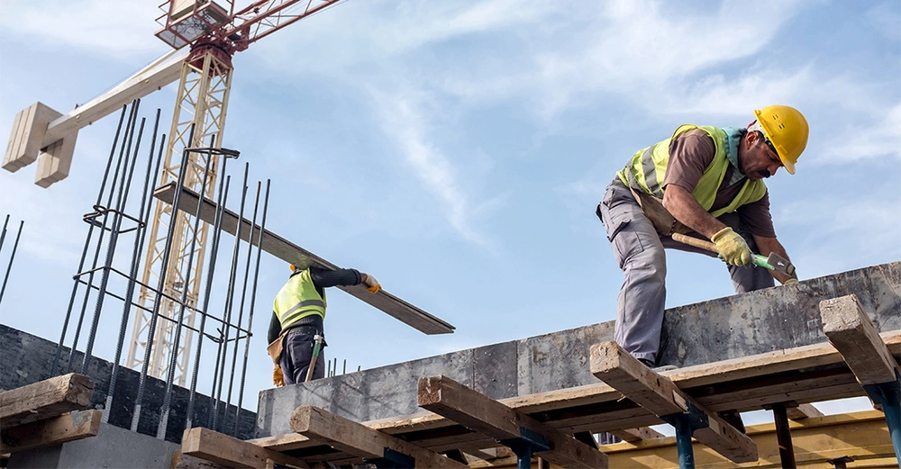 Zwei Bauarbeiter tragen Schutzhelme und Sicherheitswesten arbeiten auf einer Baustelle der eine hebt ein Holzbrett der andere hämmert auf eine Betonform im Hintergrund befindet sich ein Kran.