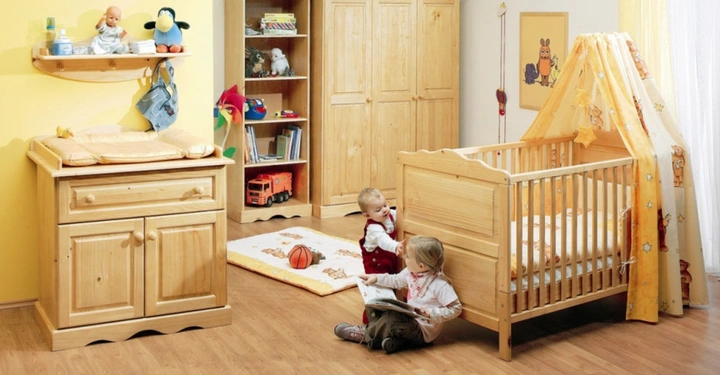 Galerie-Bild 3: Kleines Kind sitzt auf dem Boden und liest ein Buch neben einem Holzgitterbett in einem farbenfrohen Kinderzimmer mit gelbem Wickeltisch, Spielzeugregal und Schrank auf Holzboden.