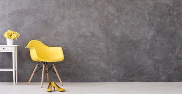 Gelber Stuhl steht auf Holzfußboden, begleitet von gelben Schuhen, daneben weißer Tisch mit gelben Blumen in einem Topf, vor einer grauen Wand.