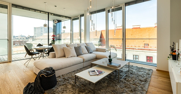 Galerie-Bild 4: Ecksofa mit Kissen steht vor großen Fenstern zusätzlich befindet sich davor ein Couchtisch mit Büchern die Umgebung zeigt eine moderne Wohnung mit Essbereich Holzboden Teppich und Blick auf Dächer.