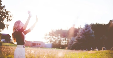Frau hebt die Arme in die Luft Steht auf einer Wiese Bäume und Häuser im Hintergrund Sonnenlicht am Himmel