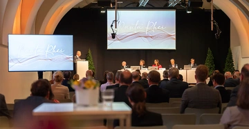 Ein Vortragender spricht am Rednerpult vor sitzendem Publikum in einer Konferenzhalle. Auf Bildschirmen ist der Text Panta Rhei zu sehen. Vier Personen sitzen an einem Podiumstisch.