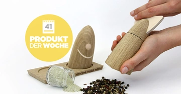 Hölzerner Pfeffermühlen-Set auf Tisch Hände mahlen Pfefferkörner daneben leeres umgekipptes Glas Text auf gelbem Hintergrund Produkt der Woche 41 attersee design edition