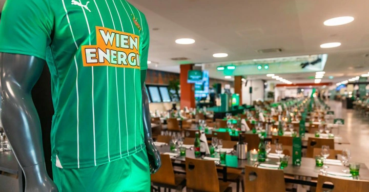 Galerie-Bild 3: Mannequin trägt grünes Fußballtrikot mit weißem Streifenmuster und dem Text WIEN ENERGIE in einem Restaurant mit vielen gedeckten Tischen und grüner Dekoration im Hintergrund.
