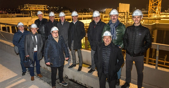 Galerie-Bild 1: Eine Gruppe von elf Männern trägt Helme und steht auf einer nächtlichen Baustelle vor einem halb fertiggestellten Gebäude mit Baustellengerüsten im Hintergrund. Auf den Helmen steht "HENN".