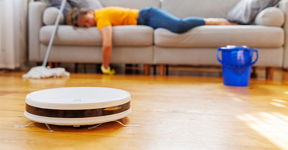 Staubsaugerroboter fährt auf Holzboden Wohnzimmer Frau liegt erschöpft auf Sofa im Hintergrund Wischmopp und blauer Eimer auf dem Boden neben ihr