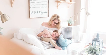 Frau sitzt auf einem weißen Sofa und umarmt lachendes Kind in heller Wohnung mit Pflanzen und Bilderrahmen an der Wand. Text im Bilderrahmen: "I hope you live a life ..." F Scott Fitzgerald