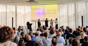Vier Redner (3 Männer, eine Frau) stehen auf Bühne vor relativ großem, sitzendem Publikum. Auf Whiteboard der Schriftzug "Living in the Glasshouse" in gelb und rosa gehalten. Im Hintergrund weiße Lamellenvorhänge, die große Glasfront verdunkeln.