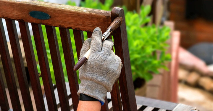 Hand streicht mit Pinsel einen Holzstuhl im Garten Kontext grünes Laub und Holzmöbel im Hintergrund