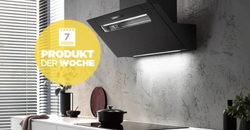 Eine schwarze Dunstabzugshaube beleuchtet eine moderne Küche mit weißen Schränken. Links befinden sich Jalousien und Pflanzen auf der Arbeitsplatte. Text: 7 PRODUKT DER WOCHE.